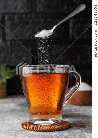 White sugar falling from teaspoon into glass cup of hot tea placed on coaster against black brick wall as background White sugar falling from teaspoon into glass cup of hot tea placed on coaster against black brick wall as background 125543051