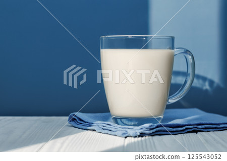 Sunlight illuminating glass milk resting on blue napkin on white wooden table with blue background, creating cozy scene 125543052