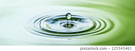 Single drop falling into calm water surface, creating concentric ripples and a crown splash, with green and white reflections, ideal for purity, nature, and wellness concepts 125543461