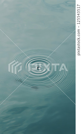 Single water droplet falling onto a calm turquoise water surface, creating expanding concentric ripples, offers a tranquil and mesmerizing view 125543517