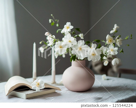 A beautiful floral arrangement of white fresh jasmine flowers and white candles on a table with an open book in a calm, cozy setting. Home decoration. This is AI-generated item 125544224