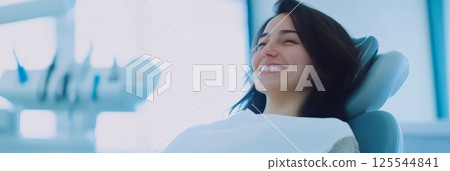 Happy female patient showing her perfect smile while sitting in a dental chair after a cosmetology treatment, enjoying the results of professional dental care in a modern clinic Happy female patient showing her perfect smile while sitting in a dental chair after a cosmetology treatment, enjoying the results of professional dental care in a modern clinic 125544841