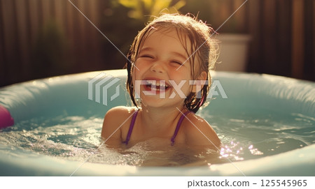 A joyful young girl with wet hair laughs happily while playing in a small inflatable pool under warm sunlight 125545965