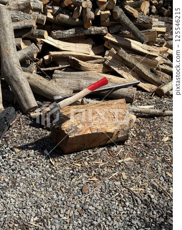 A splitting maul embedded in a log, surrounded by chopped and unchopped firewood on gravel under bright sunlight. 125546215