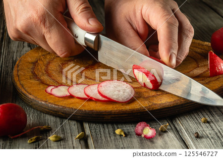 Hands skillfully slice radishes into thin rounds on a wooden cutting board. The kitchen setting displays rustic charm, highlighting the fresh ingredients being prepared 125546727