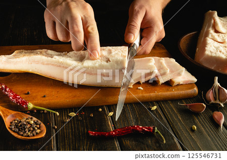Hands skillfully cut cured meat on a wooden board, surrounded by vibrant spices like garlic and chili peppers. A rustic kitchen ambiance enhances the culinary experience 125546731