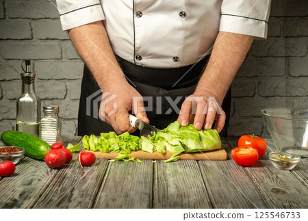 A chef skillfully chops cabbage on a wooden cutting board in a rustic kitchen. Fresh tomatoes, cucumbers, and other vegetables surround the board, enhancing the vibrant atmosphere A chef skillfully chops cabbage on a wooden cutting board in a rustic kitchen. Fresh tomatoes, cucumbers, and other vegetables surround the board, enhancing the vibrant atmosphere 125546733
