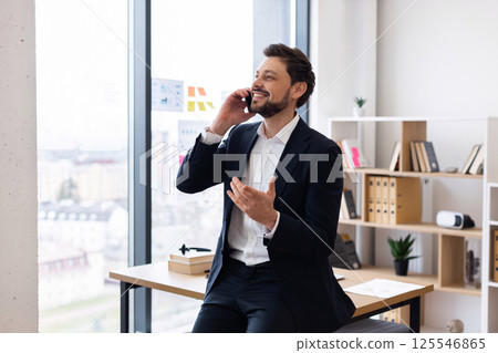 Confident Caucasian male professional in his 30s seated in modern office interior gesturing while conversing on phone, conveying positivity, technology use, communication, work discussion Confident Caucasian male professional in his 30s seated in modern office interior gesturing while conversing on phone, conveying positivity, technology use, communication, work discussion 125546865