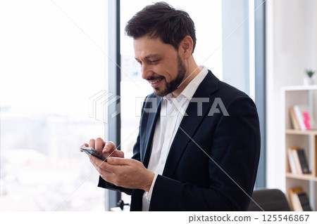 Businessman in suit using smartphone indoors during bright day. Mid-30s adult male professional in modern office setting communicating through mobile device, expressing focus. 125546867