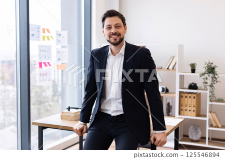 Middle-aged man wearing suit sitting on desk in well-lit modern office setting, exuding professionalism and confidence. Office surroundings include books, charts, and plants. Middle-aged man wearing suit sitting on desk in well-lit modern office setting, exuding professionalism and confidence. Office surroundings include books, charts, and plants. 125546894