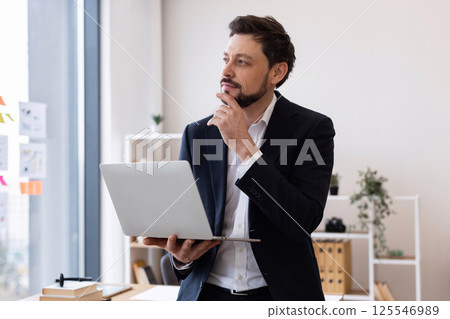 Man in early 30s wearing suit in office, holding laptop, expressing thoughtful pose, confidence. 125546989