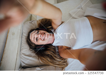 Woman in lingerie listening to music in headphones and smiling in bedroom 125547730
