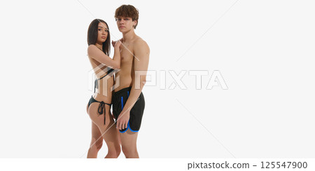 Muscular young man in black swimming trunks posing with elegant young woman in swimsuit against white background. 125547900