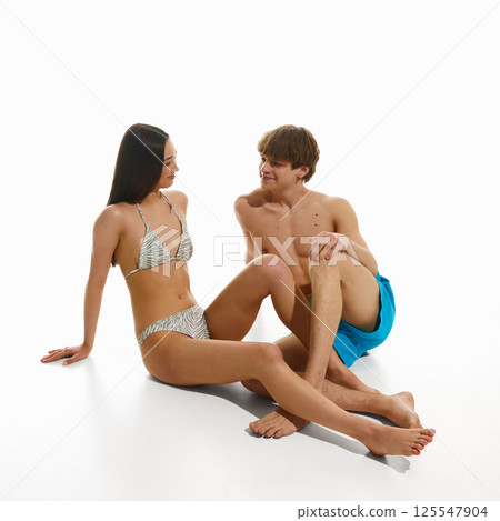 Young woman in swimsuit sitting on floor, talking to muscular shirtless man in swimming trunks against white studio background. 125547904