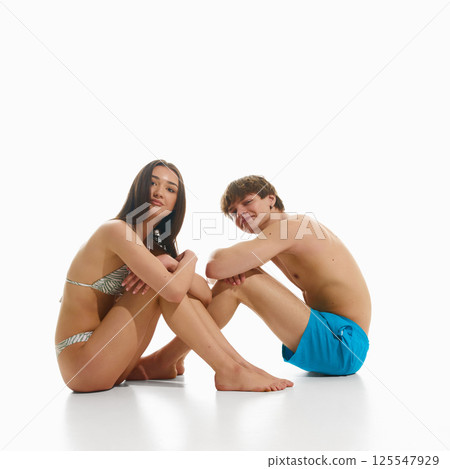 Young man and woman, both in swimwear sitting in relaxed poses, posing against white studio background. 125547929