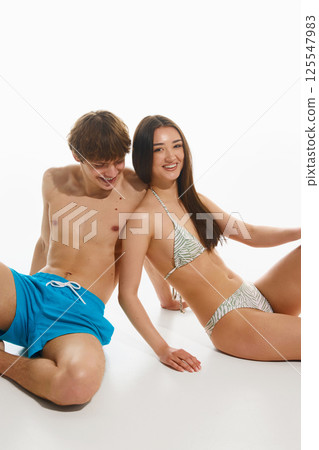 Happy, smiling young couple, fit man and slime woman, both in swimwear, posing confidently against white studio background. 125547983