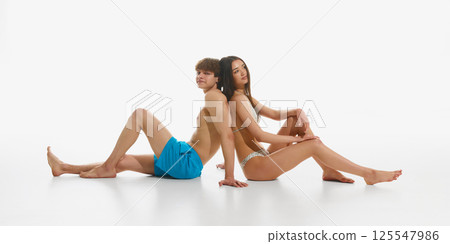 Beautiful young couple, man and woman wearing beachwear, sitting back to back, posing in relaxation against white studio background. 125547986