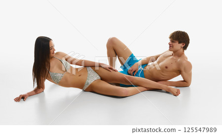 Young woman and man with fit bodies, wearing swimsuits and beach wear, posing against white studio background. Young woman and man with fit bodies, wearing swimsuits and beach wear, posing against white studio background. 125547989