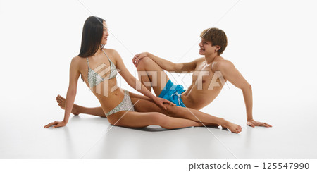 Cheerful man and woman interacting playfully, wearing stylish beach wear, posing on floor against white studio background. Cheerful man and woman interacting playfully, wearing stylish beach wear, posing on floor against white studio background. 125547990