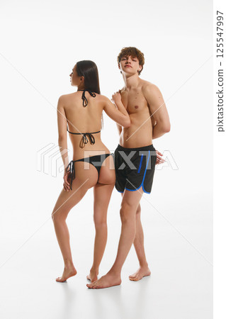 Handsome young man in black swimming trunks posing with elegant young slim woman in black swimsuit, posing against white studio background 125547997