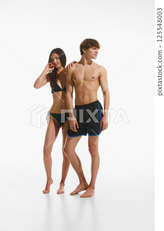 Beautiful young woman with slim, fit body in black swimsuit standing next to handsome young man with muscular body shape against white background. 125548003