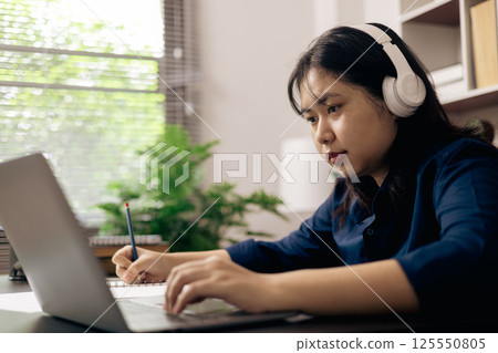 creative young student uses a laptop to complete online homework for college and university courses. Woman studies a book and conducts research for online academy using laptop in office. 125550805