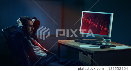 Stressed businessman sitting at desk in modern office late night, with hands behind head, looking at screen showing falling economic graphs 125552304