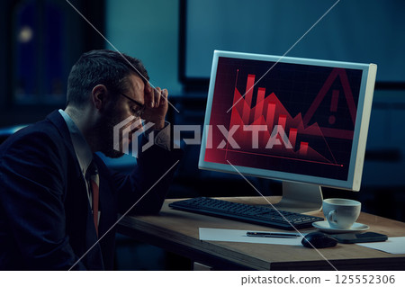 Frustrated businessman sitting by computer showing critical financial data charts, symbolizing business and financial failure. 125552306