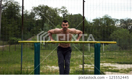 Hardy shirtless sportsman doing exercises on parallel bars at nature. Strong and muscular man working out at sports ground. Young athletic guy exercising outdoor. Concept of active healthy lifestyle 125552445