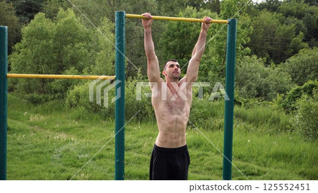 Muscular athlete doing pull ups on horizontal bar at sports ground. Young shirtless sportsman exercising at nature. Strong sporty man training outdoor. Concept of healthy and active lifestyle. Slow mo 125552451