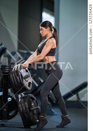 determined woman is preparing lift weights in modern fitness center. Her athletic physique highlights her dedication strength training. workout space is well equipped and inviting. 125556426