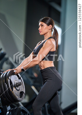 focused woman is lifting heavy weights in contemporary gym setting. She is dressed in black sports bra and leggings showcasing dedication her workout. 125556427