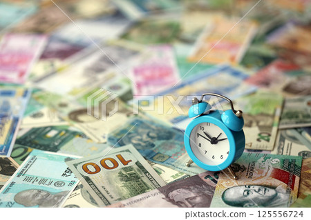 Small alarm clock on many banknotes of different currency. Background of time and money Small alarm clock on many banknotes of different currency. Background of time and money 125556724