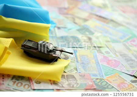Toy tank on Ukrainian flag on many banknotes of different currency. Background of war funding 125556731