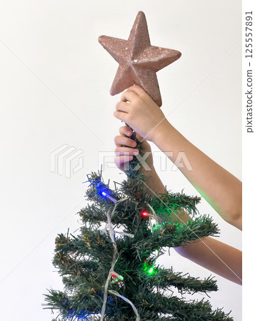Close up of child's hands placing a sparkling star on top of a decorated Christmas tree with colorful festive lights against a soft white background capturing a joyful holiday tradition Close up of child's hands placing a sparkling star on top of a decorated Christmas tree with colorful festive lights against a soft white background capturing a joyful holiday tradition 125557891