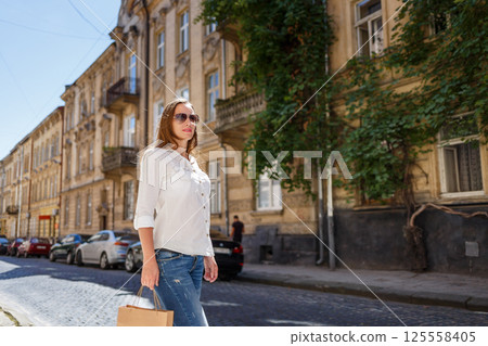 Young woman walking down a sunlit street carrying a shopping bag on a breezy day in a charming neighborhood Young woman walking down a sunlit street carrying a shopping bag on a breezy day in a charming neighborhood 125558405