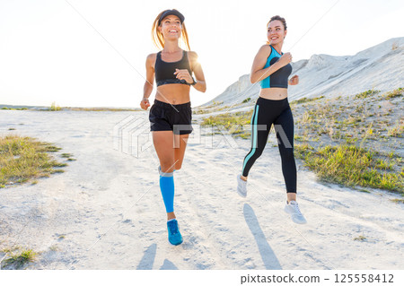 Two athletic woman enjoy outdoor running in sunlight against a scenic backdrop 125558412