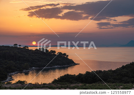The sea coast in Peljesac peninsula in southern Dalmatia in Croatia, Europe. Sunset over a tranquil bay, with a treelined coast silhouetted against the vibrant sky and reflective water. The sea coast in Peljesac peninsula in southern Dalmatia in Croatia, Europe. Sunset over a tranquil bay, with a treelined coast silhouetted against the vibrant sky and reflective water. 125558977