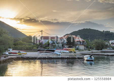 Mali Ston, village in Peljesac peninsula in Croatia, Europe. Tranquil waterfront scene with beached boats and houses bathed in soft sunlight, backed by mountains and a serene sky. 125558984