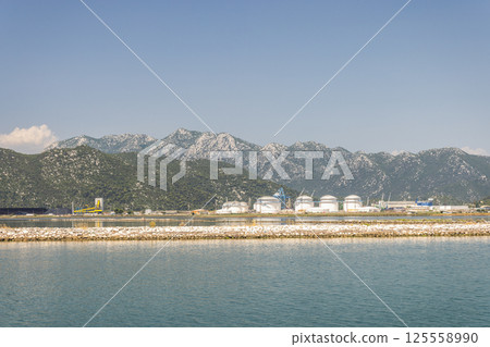 Neretva river near the mouth of river into Adriatic Sea, Croatia, Europe. Coastal Industrial Site: White storage tanks stand against mountains, under a blue sky. Calm water reflects the sunny scene. Neretva river near the mouth of river into Adriatic Sea, Croatia, Europe. Coastal Industrial Site: White storage tanks stand against mountains, under a blue sky. Calm water reflects the sunny scene. 125558990
