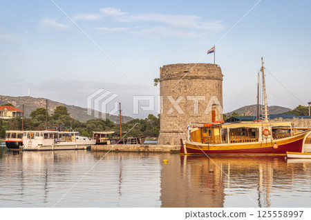 Mali Ston, village in Peljesac peninsula in Croatia, Europe. Picturesque Croatian harbor scene with boats moored near an old stone tower, reflecting in the calm water, under a soft blue sky. Mali Ston, village in Peljesac peninsula in Croatia, Europe. Picturesque Croatian harbor scene with boats moored near an old stone tower, reflecting in the calm water, under a soft blue sky. 125558997