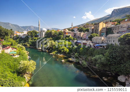 Neretva river in Mostar town, Bosnia and Herzegovina, Europe. Picturesque European cityscape featuring a river flowing through a town with historical buildings, minarets, and lush greenery. 125559011