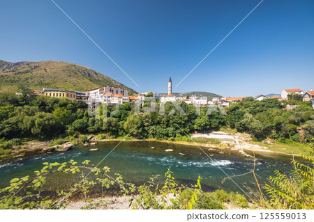 Neretva river in Mostar town, Bosnia and Herzegovina, Europe. Scenic view of a town nestled along a riverbank, featuring charming architecture and lush greenery under a clear blue sky. Peaceful. 125559013
