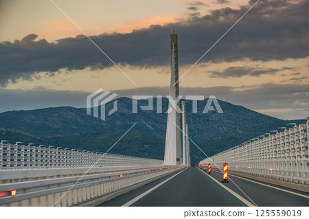 The Peljesac Bridge to Peljesac peninsula in Croatia, Europe. Driving across a modern bridge at dusk, supported by tall pillars against a backdrop of cloudy skies and distant mountains. 125559019
