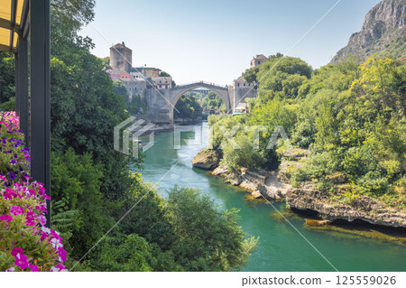 The Stari Most bridge, old bridge on Neretva river in Mostar town, Bosnia and Herzegovina, Europe. Old bridge arches across a river, flanked by historic buildings and lush greenery on a sunny day. 125559026