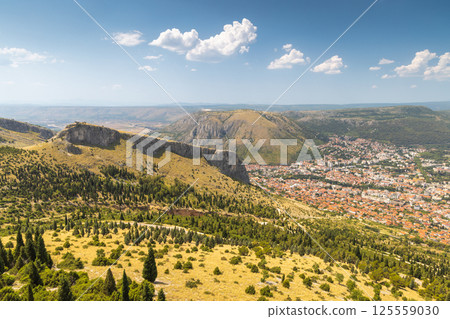 View of Mostar from the Fortica hill, Bosnia and Herzegovina, Europe. Sweeping aerial view captures a charming town nestled within a mountainous landscape under a sunny, partly cloudy sky. 125559030