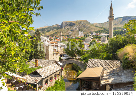 Mostar, historic town in Bosnia and Herzegovina, Europe. Picturesque old town scene with a stone bridge, tiled roofs, and a distant minaret beneath a blue sky in a European setting. 125559034