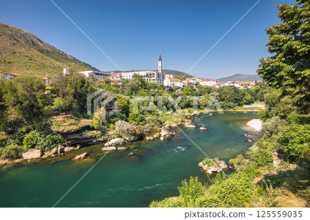 Neretva river in Mostar town, Bosnia and Herzegovina, Europe. River flows through a European town. Lush greenery surrounds buildings with mountains in background under a bright blue sky. Neretva river in Mostar town, Bosnia and Herzegovina, Europe. River flows through a European town. Lush greenery surrounds buildings with mountains in background under a bright blue sky. 125559035