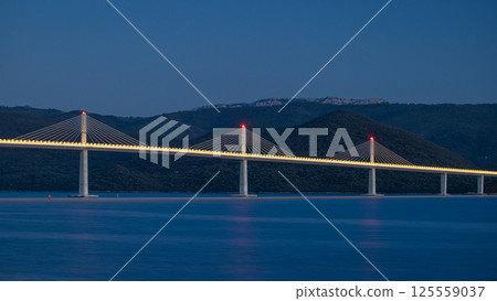 The Peljesac Bridge to Peljesac peninsula in Croatia, Europe. A modern cable-stayed bridge illuminated against a blue evening sky, spanning over calm waters with mountains in the background. 125559037