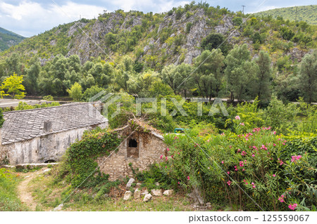 Old stone house in Blagaj village in Bosnia and Herzegovina, Europe. Overgrown stone buildings nestle among lush greenery beneath a rocky hill, evoking a sense of hidden serenity and forgotten charm. 125559067